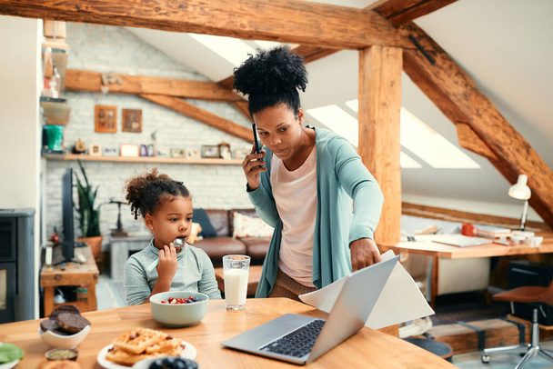 Afro-Amerikan çalışan anne cep telefonuyla konuşuyor ve yemek masasında kahvaltı ederken kızının bilgisayarını işaret ediyor.. - Fotoğraf, Görsel