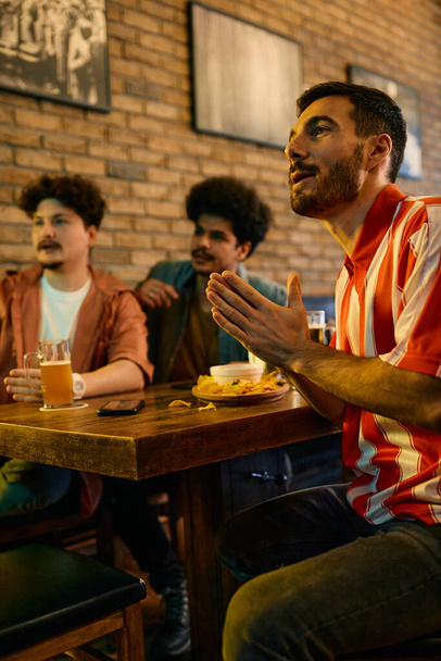 Young sports fans watching a match on TV in a pub. Focus is on man with hands clasped.  - Foto, afbeelding