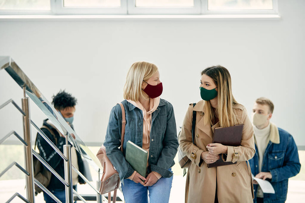 Happy students talking while walking up the stairs at university hallway and wearing protective face masks due to coronavirus pandemic. - Foto, Imagem