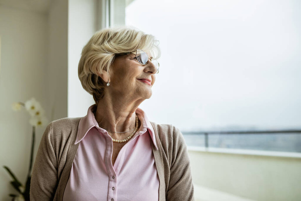 Portrait of happy mature woman relaxing by the window and looking through it. Copy space.  - Foto, afbeelding