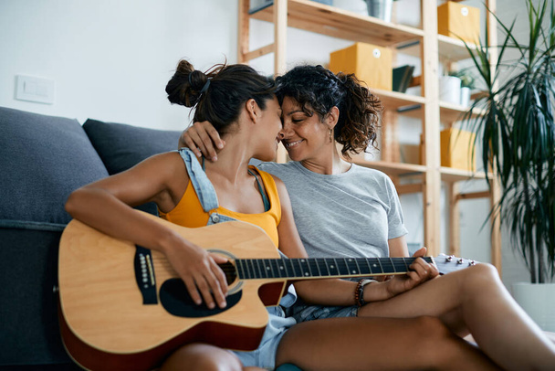 Happy female couple showing affection while playing acoustic guitar at home. - Foto, imagen