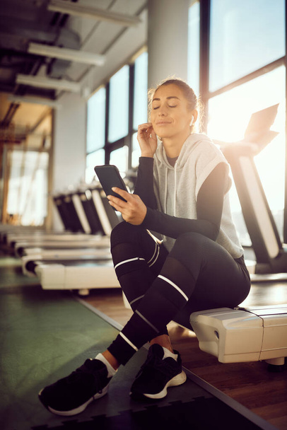 Усміхнена спортивна жінка використовує мобільний телефон і слухає музику з закритими очима, розслабляючись під час спортивних тренувань у спортзалі
.  - Фото, зображення