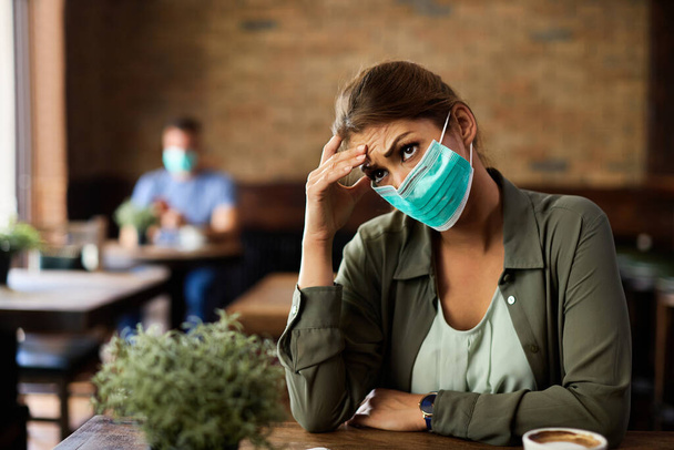 Νεαρή γυναίκα που φοράει μάσκα προσώπου και ανησυχεί για κάτι ενώ κάθεται σε ένα καφέ.,  - Φωτογραφία, εικόνα
