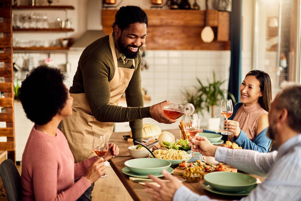 Happy black man pouring wine while having meal with his friends at dining table at home.  - Foto, immagini