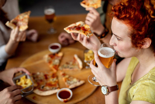 Νεαρή γυναίκα πίνει μπύρα και τρώει πίτσα ενώ είναι με τους φίλους της σε μια παμπ. - Φωτογραφία, εικόνα