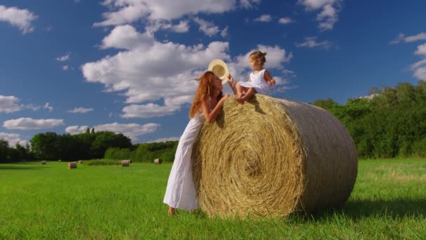 Mom and daughter in white dresses in the field. Straw stacks stack bales of hay left over from harvesting crops. Landscape of straw bales against setting sun on background. High quality 4k footage - Filmati, video