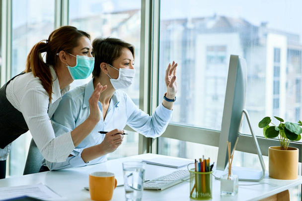 Happy businesswomen with face masks waving while making video call on desktop PC in the office during virus epidemic. - Foto, afbeelding