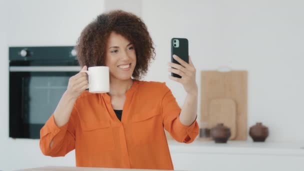 Afrikkalainen amerikkalainen tyttö bloggaaja nainen vaikuttaja tilalla kuppi teetä kahvi osoittaa kameraan tekee valokuva kirjaa video blogi online-puhelun matkapuhelin voittaa voitto reaktio web voittaa kotona keittiö - Materiaali, video