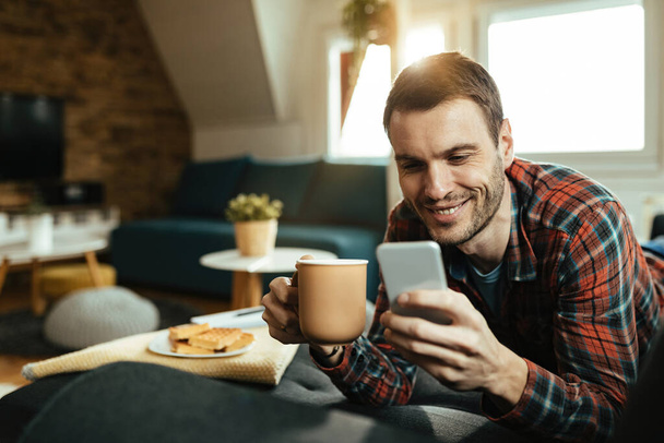 Mladý šťastný muž čtení textové zprávy na chytrém telefonu při pití kávy a relaxaci v obývacím pokoji.  - Fotografie, Obrázek