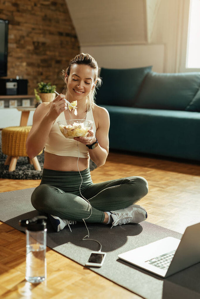 Onnellinen urheilullinen nainen istuu lattialla ja syö salaattia harjoittelun jälkeen kotona.  - Valokuva, kuva