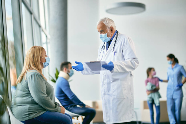 Chefarzt trägt Mundschutz, während er mit Patientin im Wartezimmer eines Krankenhauses spricht. - Foto, Bild