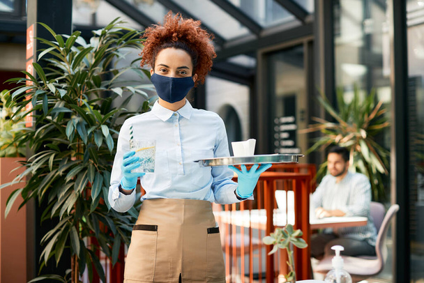 Portrait of waitress serving drinks while wearing protective face mask and gloves in a cafe and looking at camera. - Foto, Bild