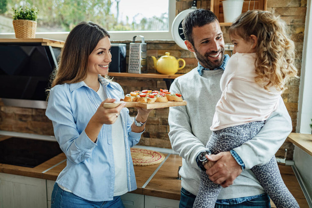 Happy parents talking with their small daughter while offering her to try bruschetta in the kitchen.  - Fotografie, Obrázek