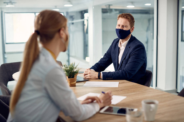 Happy entrepreneur communicating with female coworker while wearing protective face mask in the office due to coronavirus pandemic. - Foto, Imagen