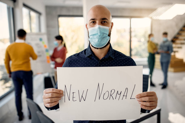 Businessman with face mask holding placard with 'new normal' inscription while working in the office during COVID-19 pandemic. - Foto, imagen