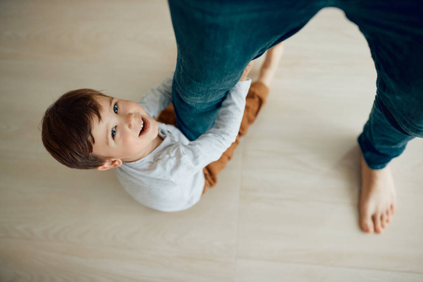 High angle view of carefree kid having fun while holding on to his fathers leg at home and looking at camera.   - Foto, immagini