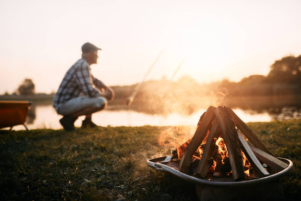 Close-up of bonfire with man fishing on the river in the background at sunset. Copy space. - 写真・画像