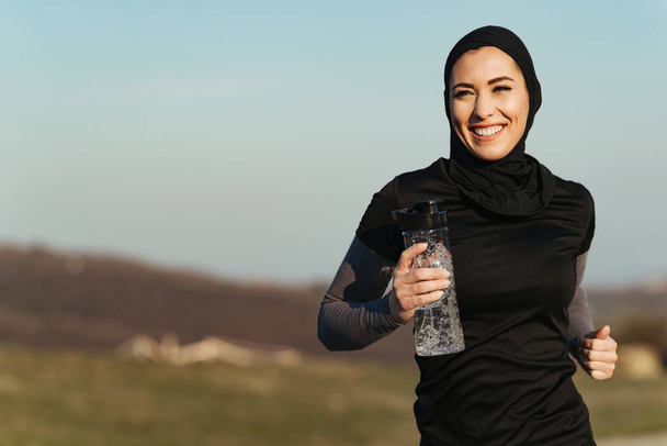 Молода щаслива атлетична жінка, що несе пляшку води під час бігу на природі
.  - Фото, зображення