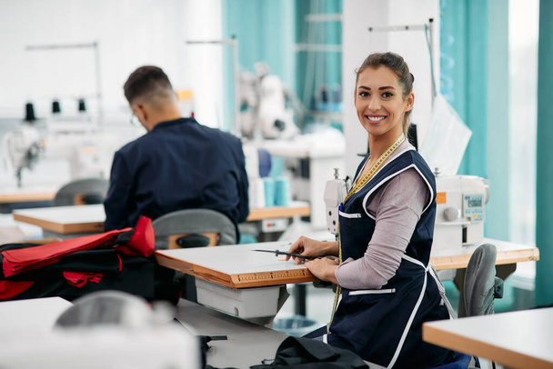 Happy seamstress sewing at production line in textile factory and looking at camera. - Foto, Bild