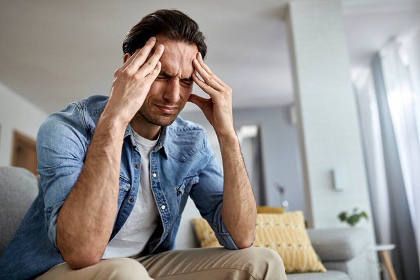 Low angle view of distraught man holding his head in pain while sitting in the living room.  - Photo, Image