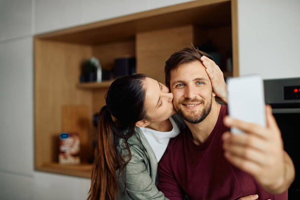 Affectionate woman kissing her husband while he is taking selfie with mobile phone at home. - Foto, Imagen
