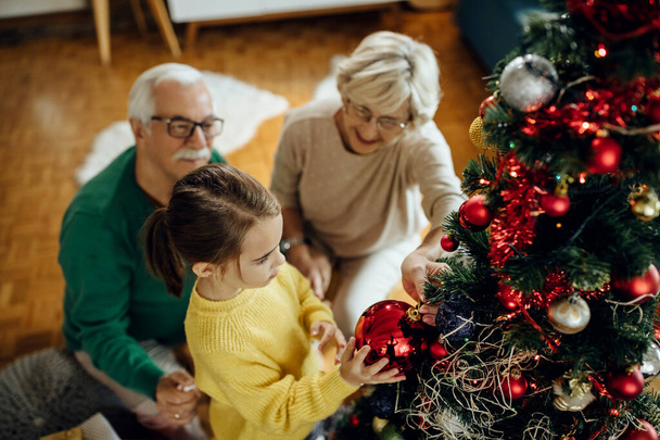 Korkea kulma näkymä pikkutyttö ja hänen isovanhemmat koristelu joulukuusi kotona.  - Valokuva, kuva