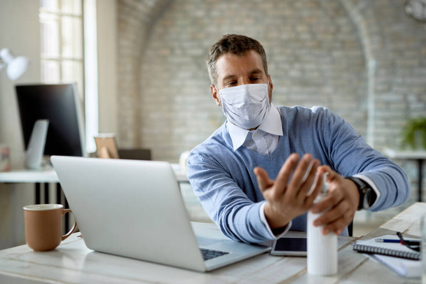 Businessman with protective face mask disinfecting his hands while working in the office.  - Foto, Bild