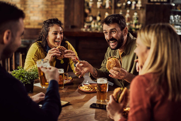 Молодий щасливий чоловік розважається з друзями під час їжі гамбургерів і пива в пабі
. - Фото, зображення