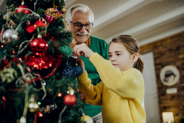 Small girl decorating Christmas tree with her grandfather in the living room.  - Foto, Imagen