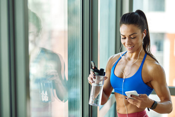 Young muscular build woman drinking water and using smart phone by the window.  - 写真・画像