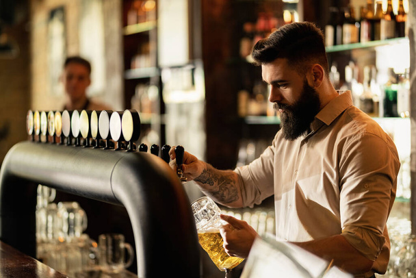 Young bartender pouring draft beer in beer glass while working at bar counter.  - Zdjęcie, obraz