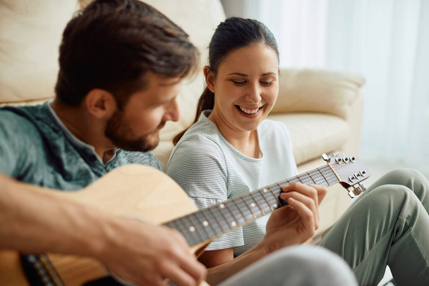 Young happy woman listening to her husband who is playing acoustic guitar at home. - Foto, imagen