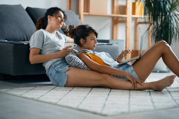 Nuori nainen ja hänen tyttöystävänsä käyttävät kannettavaa tietokonetta rentoutuessaan lattialla kotona.  - Valokuva, kuva