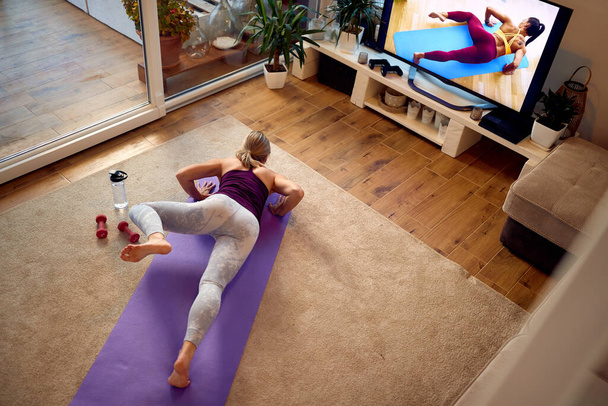 High angle view of athletic woman watching exercises class on a TV and practicing alligator push-ups in the living room. - Foto, immagini