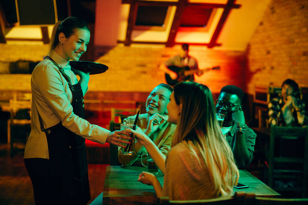 Jonge vrolijke serveerster serveert drankjes aan een groep mensen in een bar 's nachts.  - Foto, afbeelding