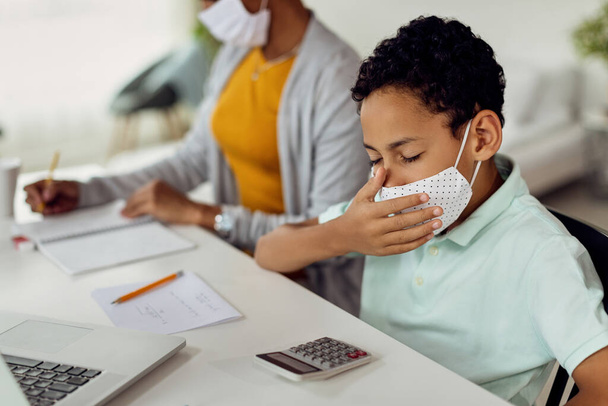 Small black boy wearing protective face mask and yawning while studying at home due to COIVD-19 epidemic.  - Foto, afbeelding