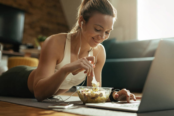 Young smiling sportswoman using laptop and eating salad while lying down on floor.  - Foto, Bild