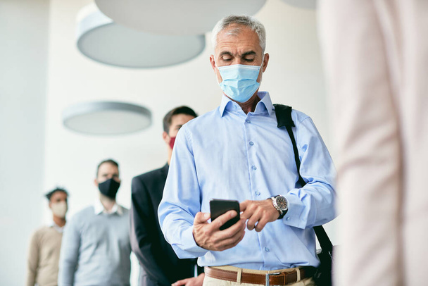 Mature businessman text messaging on mobile phone while standing in line and wearing protective face mask due to coronavirus pandemic.  - Foto, imagen