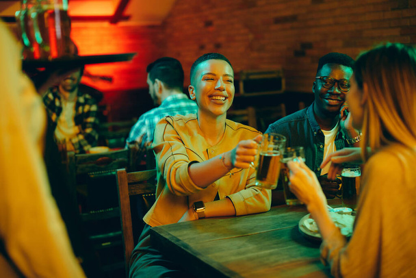 Група молодих щасливих людей готує пиво і розважається вночі в пабі. Фокус на чорній жінці
.  - Фото, зображення