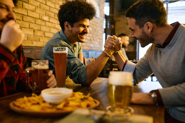 Happy Lebanese man and his male friend arm wrestling and having fun while drinking beer in a pub.  - Foto, Imagem