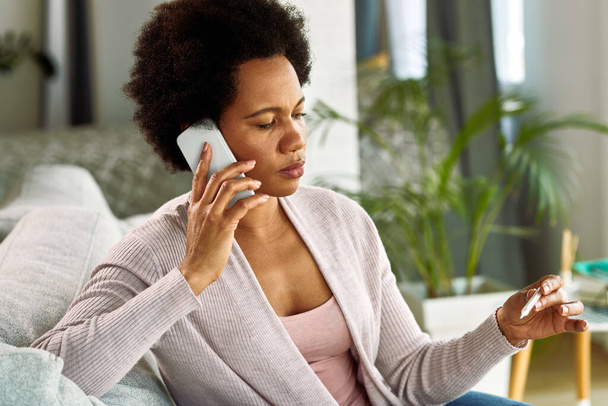 Zwarte vrouw controleren van haar temperatuur met een thermometer tijdens het communiceren op mobiele telefoon thuis.  - Foto, afbeelding