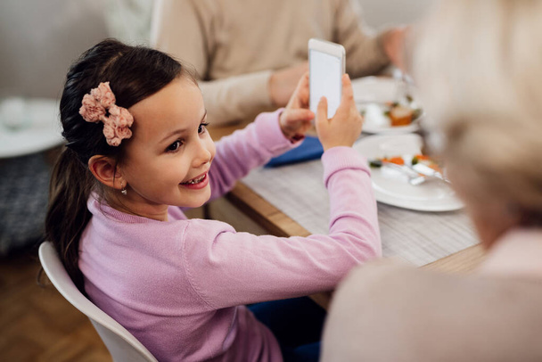 Happy small girl using mobile phone and talking to her grandmother during lunch time at dining table.  - Foto, afbeelding