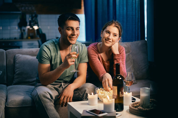 Smiling couple drinking wine and eating popcorn while watching movie on TV at night at home.  - Photo, image