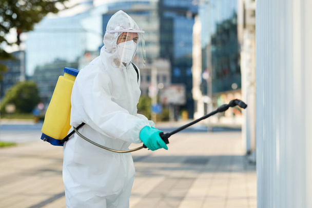 Медицинский работник в защитном костюме дезинфицирует городскую территорию с целью предотвращения распространения эпидемии коронавируса.  - Фото, изображение