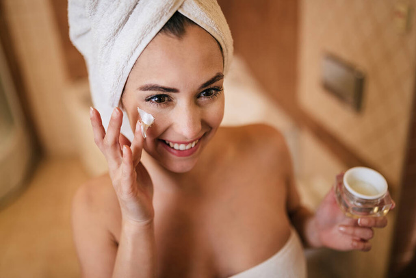 Young beautiful woman wrapped in towel applying moisturizer on her face in the bathroom. - Foto, imagen
