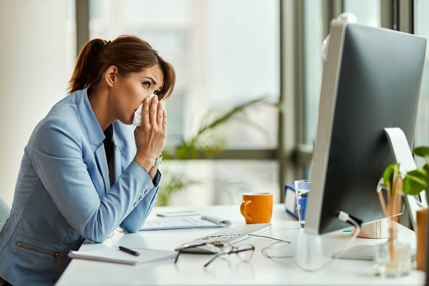 Young entrepreneur sneezing in a tissue while working at her office desk.  - Photo, Image