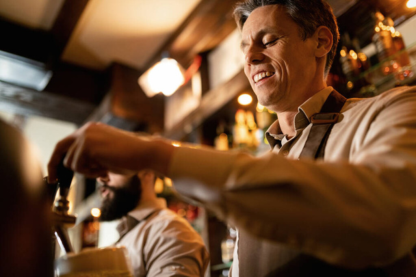Low angle view of happy barista pouring draft beer while working in a pub.  - Foto, imagen