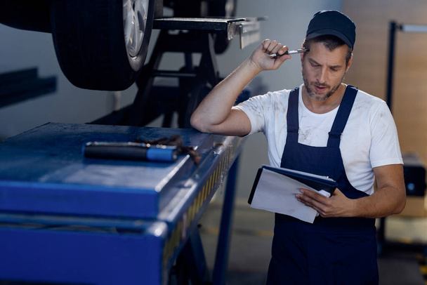 Pensive mechanic reading paperwork while examining car in auto repair shop.  - Photo, image
