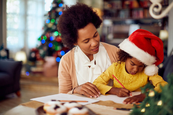 African American mother enjoying on Christmas with her daughter who is drawing something on a piece of paper at home. - Foto, Imagem