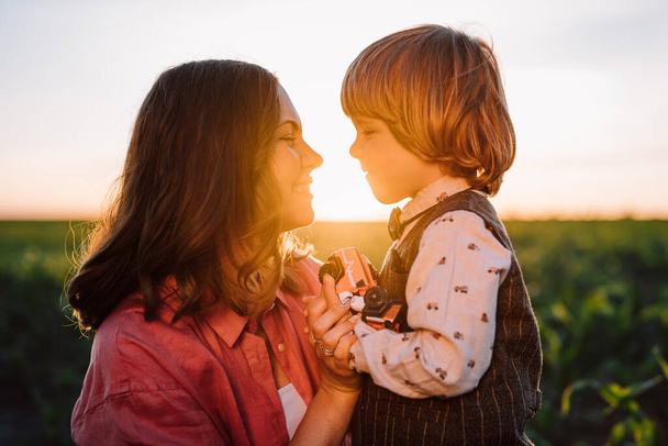 Tender scene of loving son with mom on sunset backdrop. Beautiful family. Cute 3 year old kid with mother. Parenthood, childhood, happiness, children wellbeing concept. High quality photo - Photo, image